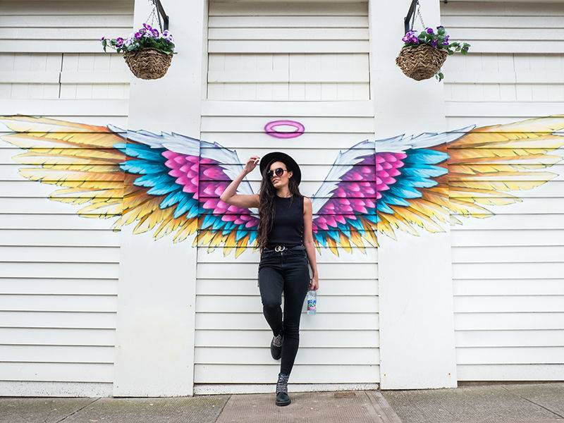 A woman in front of angel-wing graffiti, holding a bottle of macb