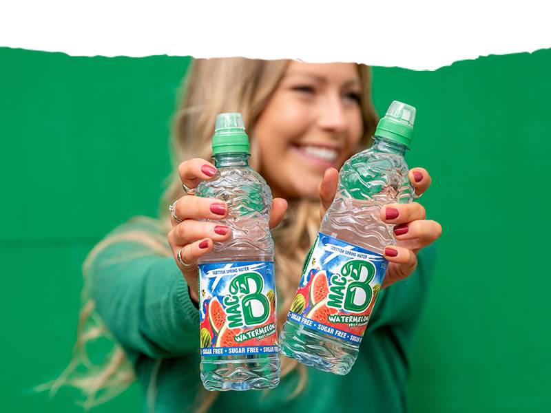 A woman with a green jumper holding two bottles of Macb watermelon flavoured spring water
