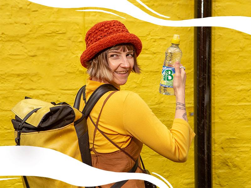 A girl in yellow clothing in front of a yellow wall, holding a bottle of Macb and smiling